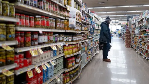 رجل يقف في ممر سوبرماركت ويحمل سلة تسوق، محاط بمختلف الأطعمة المعلبة والمنتجات، في سياق أزمة أسعار المواد الغذائية.