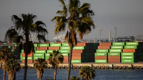 حاويات شحن تحمل علامة "إيفر غرين" في ميناء، محاطة بأشجار النخيل، تعكس النشاط التجاري في ظل التوترات التجارية العالمية.