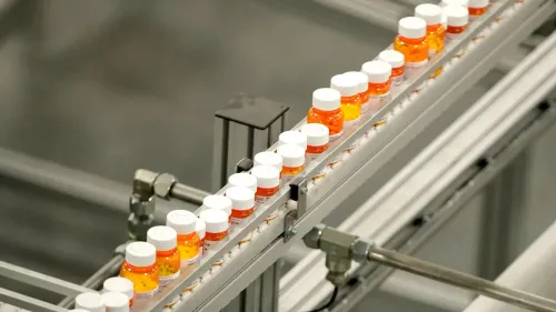 زجاجات أدوية ملونة مرتبة على حزام ناقل في مصنع، تعكس جهود الإدارة الأمريكية للحد من أسعار الأدوية في السوق.