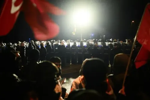 مظاهرة ليلية حاشدة، حيث يتجمع المتظاهرون أمام صف من قوات الشرطة المجهزة بمعدات مكافحة الشغب، مع وجود علم في المقدمة.