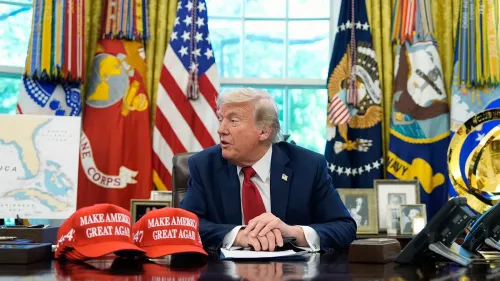 ترامب يتحدث في المكتب البيضاوي بينما تظهر قبعات "اجعل أمريكا عظيمة مرة أخرى" على الطاولة، وسط أعلام وطنية وخرائط.