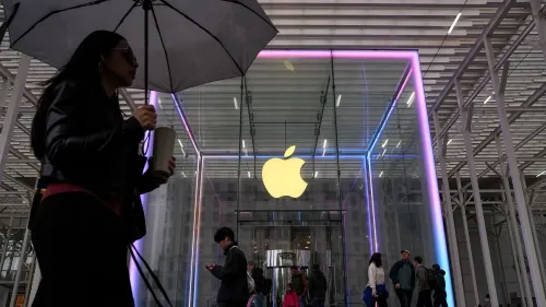 امرأة تحمل مظلة واقفة أمام متجر Apple الذي يتميز بشعار الشركة المضيء، مع وجود زبائن آخرين في الخلفية.