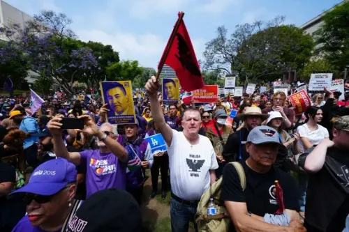 تجمع حاشد في لوس أنجلوس يتضمن متظاهرين يحملون لافتات وشعارات ضد قوانين الهجرة، مع وجود علم أحمر في المقدمة.