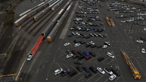 صورة جوية لموقف سيارات واسع يحتوي على العديد من السيارات، مع وجود قطارات شحن في الخلفية، تعكس تأثير الرسوم الجمركية على صناعة السيارات.