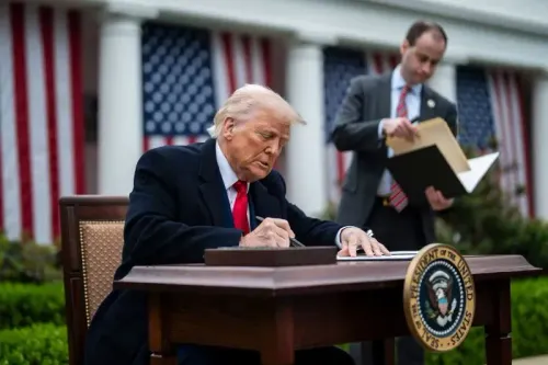 ترامب يوقع وثيقة في حديقة البيت الأبيض، مع وجود علم أمريكي خلفه، وسط أجواء تعكس التوترات الاقتصادية والسياسية.
