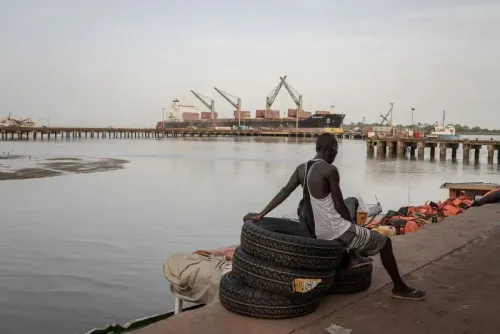 رجل يجلس على إطارات قديمة بجانب رصيف مائي، مع سفن شحن كبيرة في الخلفية، مما يعكس النشاط التجاري في المنطقة.