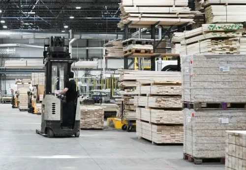 مستودع يحتوي على كميات كبيرة من الخشب، مع عامل يقود رافعة شوكية بين الرفوف، مما يعكس تأثير التعريفات الجمركية على الصناعة.