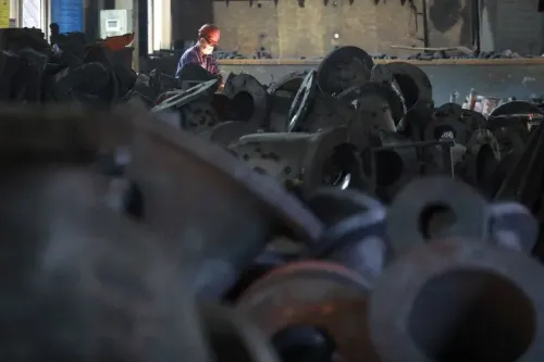 عامل يرتدي خوذة وقناعًا في مصنع، محاط بأجزاء معدنية كبيرة، يمثل تأثير السياسات الاقتصادية على التصنيع الأمريكي.