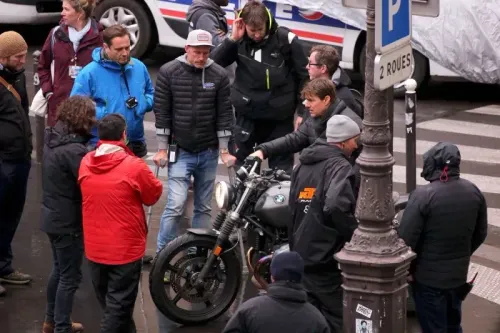 مجموعة من العاملين في صناعة السينما تتجمع حول دراجة نارية أثناء تصوير مشهد في موقع خارجي، مما يعكس التعاون الدولي في إنتاج الأفلام.