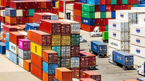 حاويات شحن ملونة مكدسة في ميناء، مع شاحنات تسليم تتحرك بينها، تعكس تأثير التعريفات الجمركية الجديدة على التجارة العالمية.