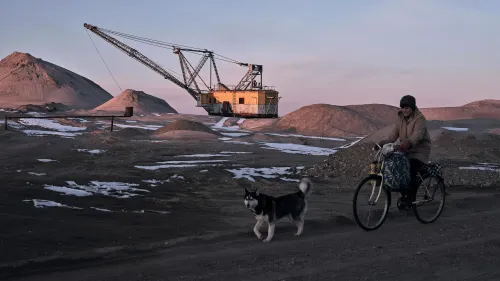 امرأة تركب دراجة قرب موقع تعدين، مع وجود كلب يرافقها، وآلة حفر كبيرة في الخلفية، تعكس النشاط الصناعي في المنطقة.