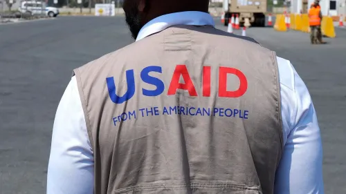 موظف من الوكالة الأمريكية للتنمية الدولية (USAID) يرتدي سترة تحمل شعار الوكالة، في موقع عمل خارجي، مع خلفية تظهر منطقة مفتوحة.