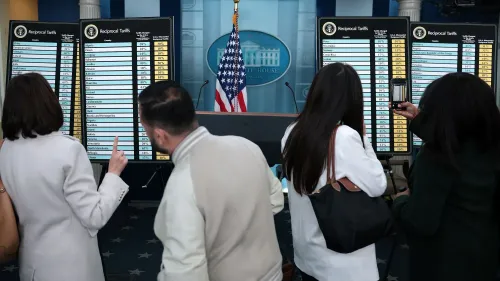 شخصان يتفقدان لوحات تعرض معدلات الرسوم الجمركية المتبادلة في مؤتمر صحفي بالبيت الأبيض، مع العلم الأمريكي خلفهم.