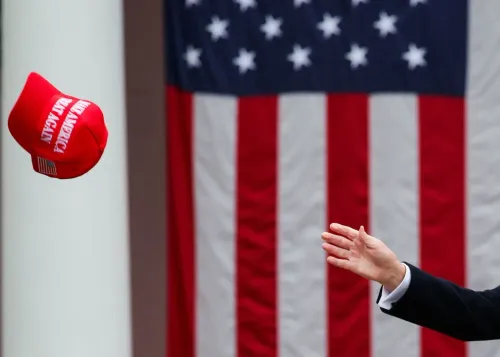 قبعة حمراء مكتوب عليها "اجعل أمريكا عظيمة مرة أخرى" تُلقى في الهواء أمام علم أمريكي، تعكس أجواء السياسة الأمريكية الحالية.