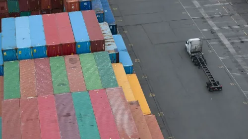 صورة لميناء شحن يظهر حاويات ملونة مرتبة بشكل منظم وشاحنة نقل تتحرك في المساحة الفارغة، تعكس تأثير الحرب التجارية على الاقتصاد الأمريكي.