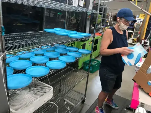 امرأة ترتدي قناعًا تعمل في مصنع، حيث تقوم بتعبئة ألعاب الحيوانات الأليفة، مع وجود أطباق زرقاء مرتبة على رفوف خلفها.