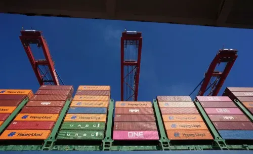 حاويات شحن ملونة في ميناء مع رافعات تحميل تحت سماء زرقاء، تعكس تأثير التعريفات الجمركية الأمريكية على التجارة العالمية.