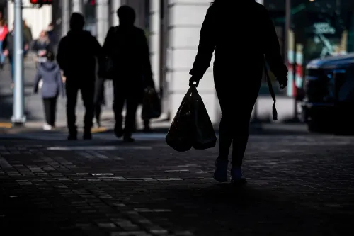 امرأة تحمل حقيبة تسوق تمشي في الشارع، بينما تظهر أشخاص آخرون في الخلفية. تعكس الصورة تأثير التعريفات الجمركية على الأسر الأمريكية.