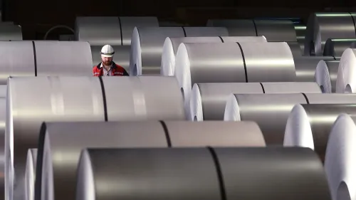 عامل يرتدي خوذة يعمل وسط لفات من الألومنيوم في مصنع، مما يعكس تأثير التعريفات الجمركية على الصناعة الأمريكية.