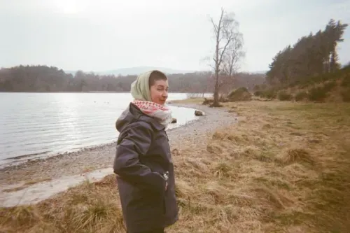 امرأة ترتدي حجابًا وتبتسم، تقف على ضفة بحيرة محاطة بالطبيعة، تعكس روح الأمل في سياق الإضراب عن الطعام.