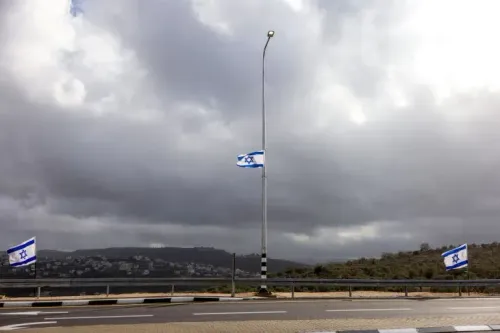 علم إسرائيلي يرفرف على عمود إنارة في منطقة جبلية تحت سماء غائمة، مع مشهد لقرية في الخلفية.