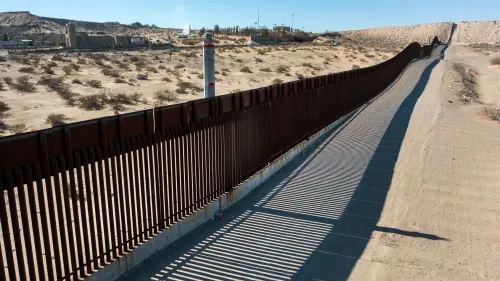 سياج الحدودي بين الولايات المتحدة والمكسيك في منطقة صحراوية، مع تفاصيل عن الطريق المجاور وظلاله، يعكس جهود تأمين الحدود الجنوبية.