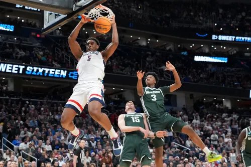 لاعب كرة سلة من جامعة كونيتيكت يقوم بعملية دنك مثيرة خلال مباراة NCAA، مع وجود لاعبين من الفريق المنافس في الخلفية.