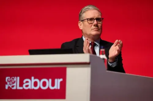 كير ستارمر، زعيم حزب العمال البريطاني، يظهر في مؤتمر صحفي، مع خلفية حمراء وشعار الحزب. يواجه تحديات سياسية كبيرة.