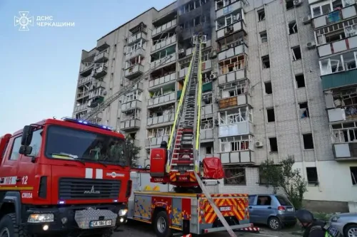 عربة إطفاء أمام مبنى سكني متضرر، حيث تتصاعد أدخنة من الطوابق العليا بعد هجوم صاروخي، مع وجود فرق الإنقاذ في الموقع.