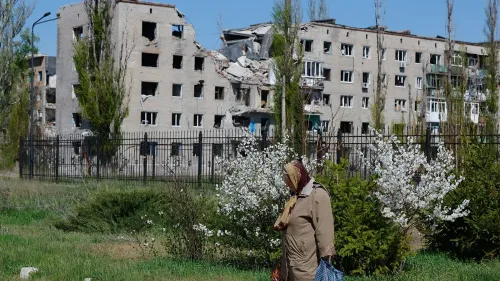 امرأة مسنّة تسير بجوار مبنى مدمر في أوكرانيا، حيث تظهر آثار الدمار الناجم عن الهجمات، مع زهور بيضاء تتفتح في المقدمة.