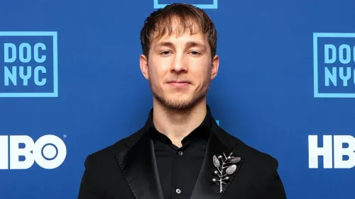 رجل يرتدي بدلة سوداء مع دبوس زينة، يقف أمام خلفية زرقاء تحمل شعارات \"DOC NYC\" و\"HBO\"، يبدو مبتسمًا وواثقًا.