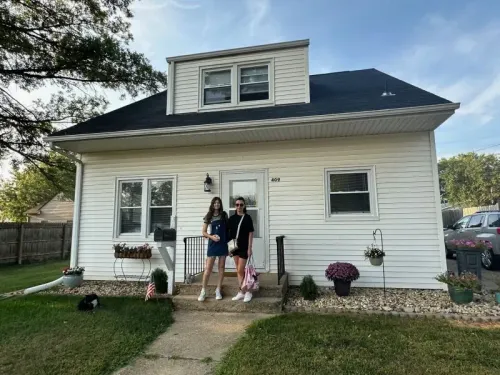 امرأتان أوكرانيتان واقفتان أمام منزل أبيض في هارتسفيل، حيث تعكسان تحديات إعادة بناء حياتهما بعد الهجرة إلى الولايات المتحدة.