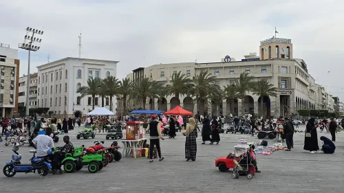 ساحة مزدحمة في طرابلس مع عائلات وأطفال يلعبون، بينما تظهر المباني التاريخية والنخيل في الخلفية.