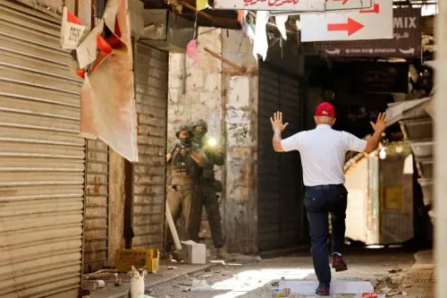 رجل فلسطيني يرتدي قبعة حمراء يرفع يديه في زقاق ضيق بنابلس، بينما يواجه مجموعة من الجنود الإسرائيليين المسلحين.