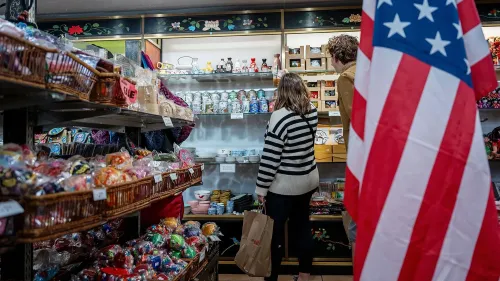امرأة ورجل يتسوقان في متجر مليء بالهدايا التذكارية، مع العلم الأمريكي في المقدمة، مما يعكس تأثير الاقتصاد الأمريكي على سلوك المستهلكين.