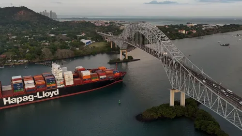 سفينة حاويات تابعة لشركة "Hapag-Lloyd" تعبر قناة بنما تحت جسر معلق، مع منظر للمدينة في الخلفية، تعكس أهمية القناة في التجارة العالمية.