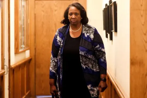 امرأة تسير في ممر، ترتدي ملابس أنيقة مع معطف مزخرف، تعكس أجواء التوتر السياسي حول إقالتها من مجلس الاحتياطي الفيدرالي.