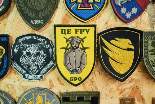 شعارات عسكرية متنوعة على خلفية خشبية، تتضمن رموزًا تعبر عن وحدات مختلفة، مع التركيز على شعار يحمل صورة شخص يتحكم بطائرة مسيرة.