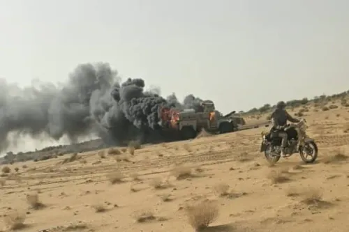 دخان أسود يتصاعد من مركبة محترقة في صحراء النيجر مع رجل على دراجة نارية، يعكس تصاعد العنف والجماعات المسلحة في الساحل.