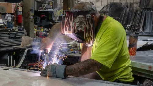 عامل يقوم باللحام في ورشة عمل، مع انبعاث شرارات ودخان، مما يعكس أهمية الصناعة في ظل ارتفاع أسعار الجملة والتضخم.