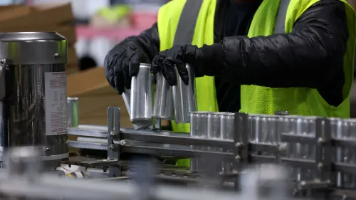 عامل يرتدي سترة عاكسة وقفازات، يقوم بتجميع علب معدنية على خط إنتاج، في سياق انخفاض أسعار الجملة في الولايات المتحدة.