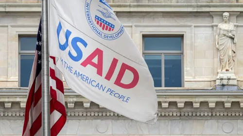 علم الوكالة الأمريكية للتنمية الدولية (USAID) يتدلى بجانب العلم الأمريكي، مع واجهة مبنى تاريخي في الخلفية، مما يعكس تأثير الوكالة في تقديم المساعدات الإنسانية.