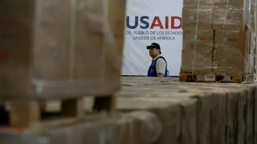 موظف في الوكالة الأمريكية للتنمية الدولية (USAID) يقف أمام لافتة الوكالة وسط صناديق مساعدات، في سياق مناقشات حول مستقبل الوكالة.