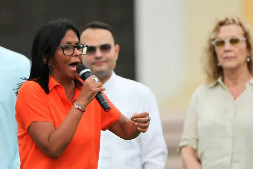 تظهر الصورة Delcy Rodríguez، رئيسة فنزويلا بالإنابة، تتحدث في تجمع عام، مرتدية قميصًا برتقاليًا، مع وجود شخصين خلفها.
