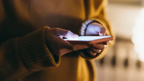 يد تحمل هاتفًا ذكيًا مضاءً في غرفة مظلمة، مما يعكس القلق حول الصور غير المرغوب فيها التي تم إنشاؤها بالذكاء الاصطناعي.