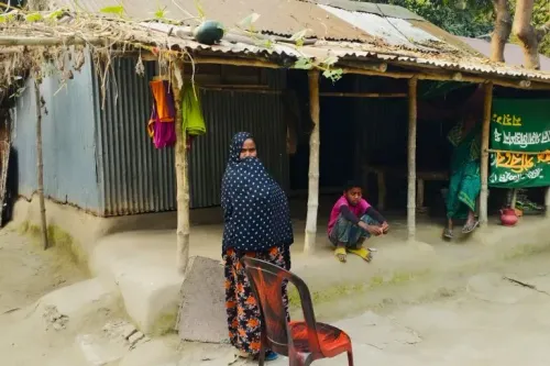 امرأة ترتدي الحجاب تقف أمام منزل بسيط في قرية بايغوني، بينما يجلس طفل بجوارها، مما يعكس التحديات الاجتماعية والاقتصادية في المنطقة.
