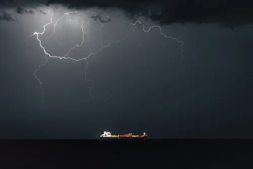 سفينة شحن تبحر في البحر تحت سماء ملبدة بالغيوم، مع وميض البرق في الخلفية، مما يعكس حالة عدم الاستقرار في مضيق هرمز.