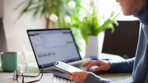 امرأة تستخدم هاتفها الذكي أثناء العمل على الكمبيوتر المحمول في بيئة منزلية مريحة، مع وجود نباتات في الخلفية.