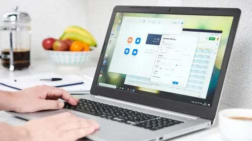 صورة تظهر يد شخص تستخدم الكمبيوتر المحمول لجدولة اجتماع عبر تطبيق زووم، مع خلفية تحتوي على فواكه ومشروبات.
