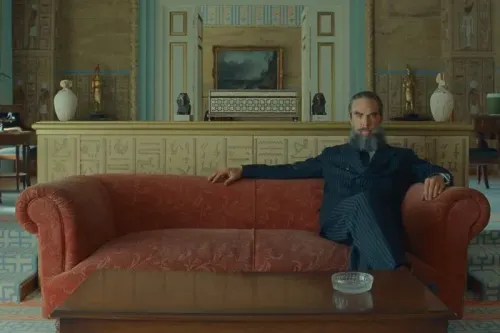 مشهد من فيلم "المخطط الفينيقي" لويس أندرسون، يظهر شخصية رجل أعمال أنيق يجلس على أريكة حمراء في بيئة فاخرة.